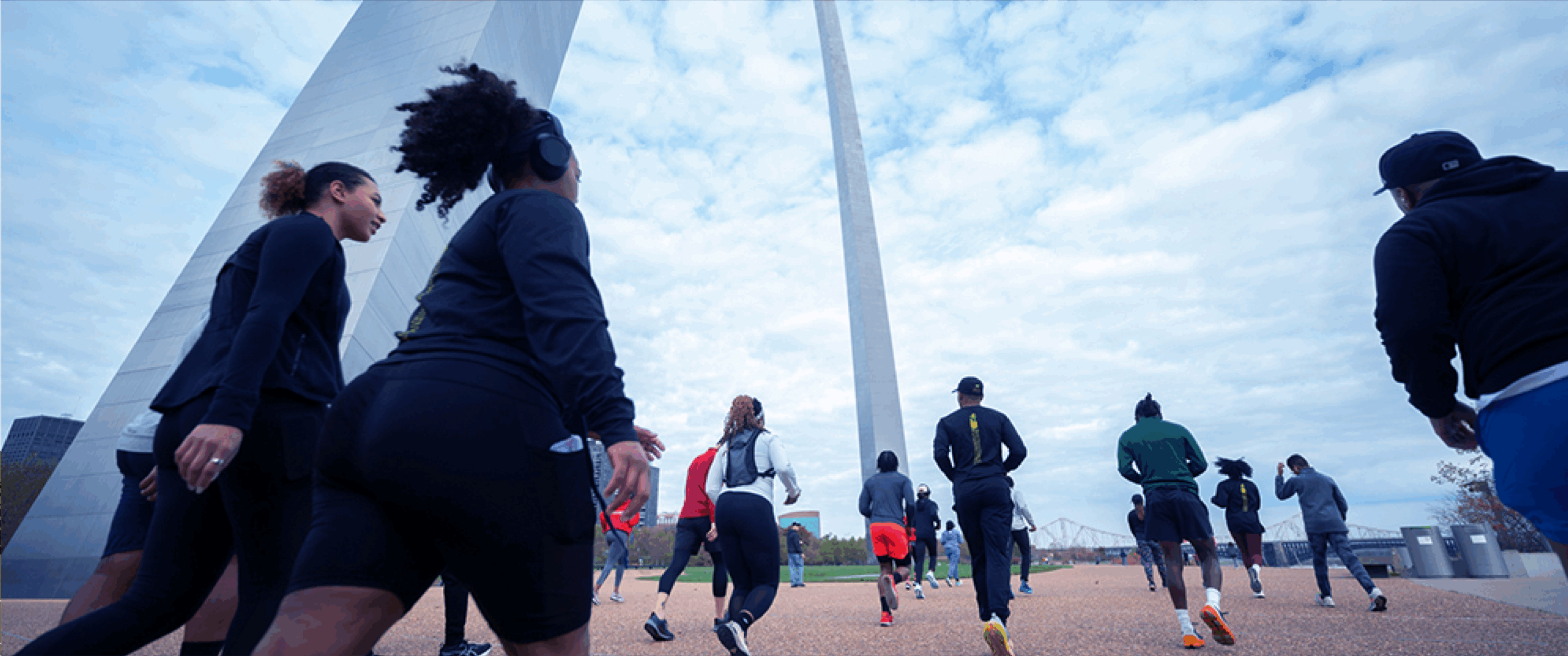A group of runners in athletic clothing jog away from the camera toward a towering stainless-steel arch under a cloudy sky, captured from a low angle with the monument rising overhead.