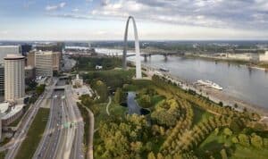 Gateway Arch rising beside curving Mississippi River, riverside park with winding paths, adjacent highway and city skyline.