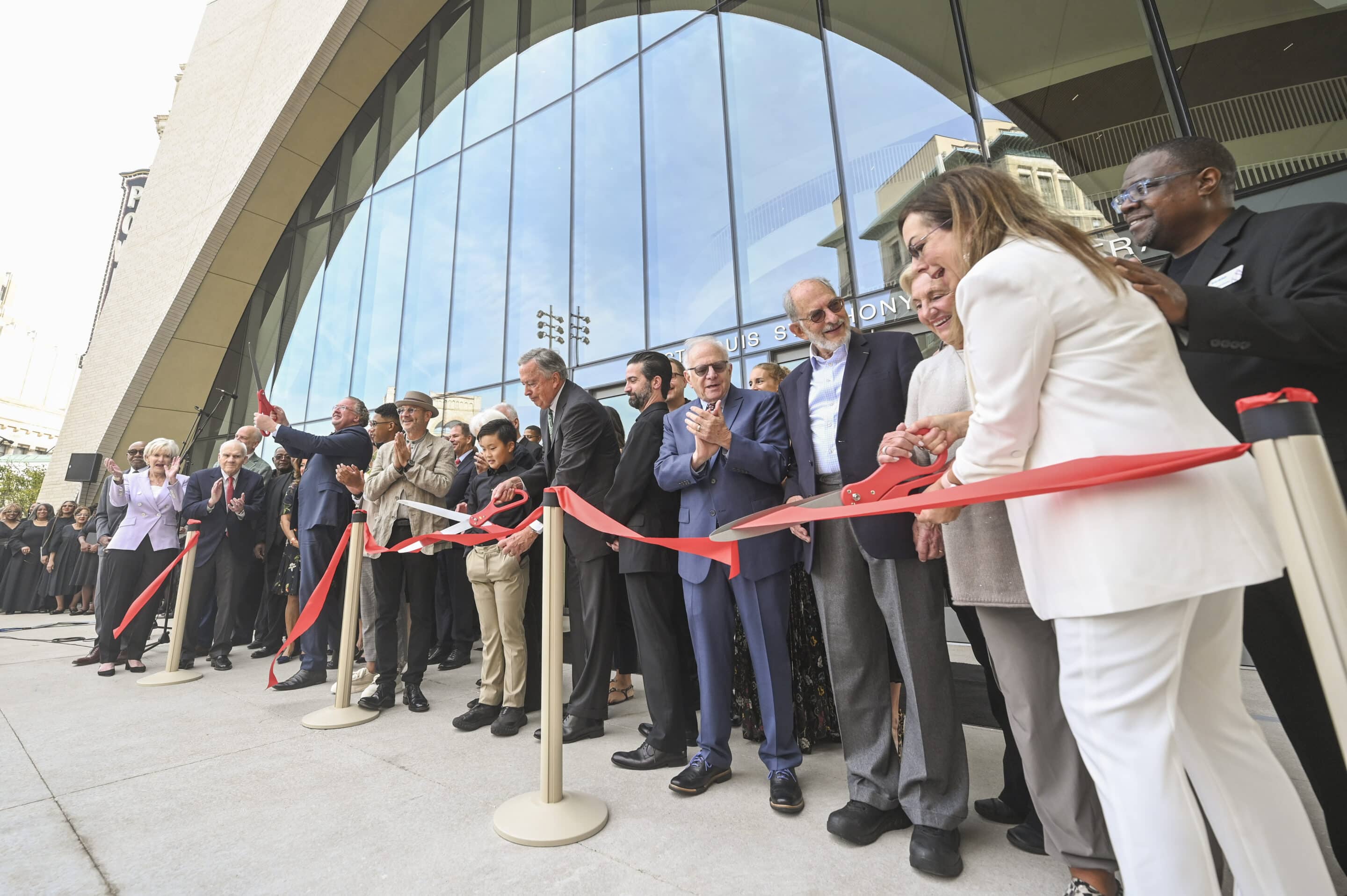 Officials and guests cut a long red ribbon with oversized scissors in front of a curved glass building entrance.