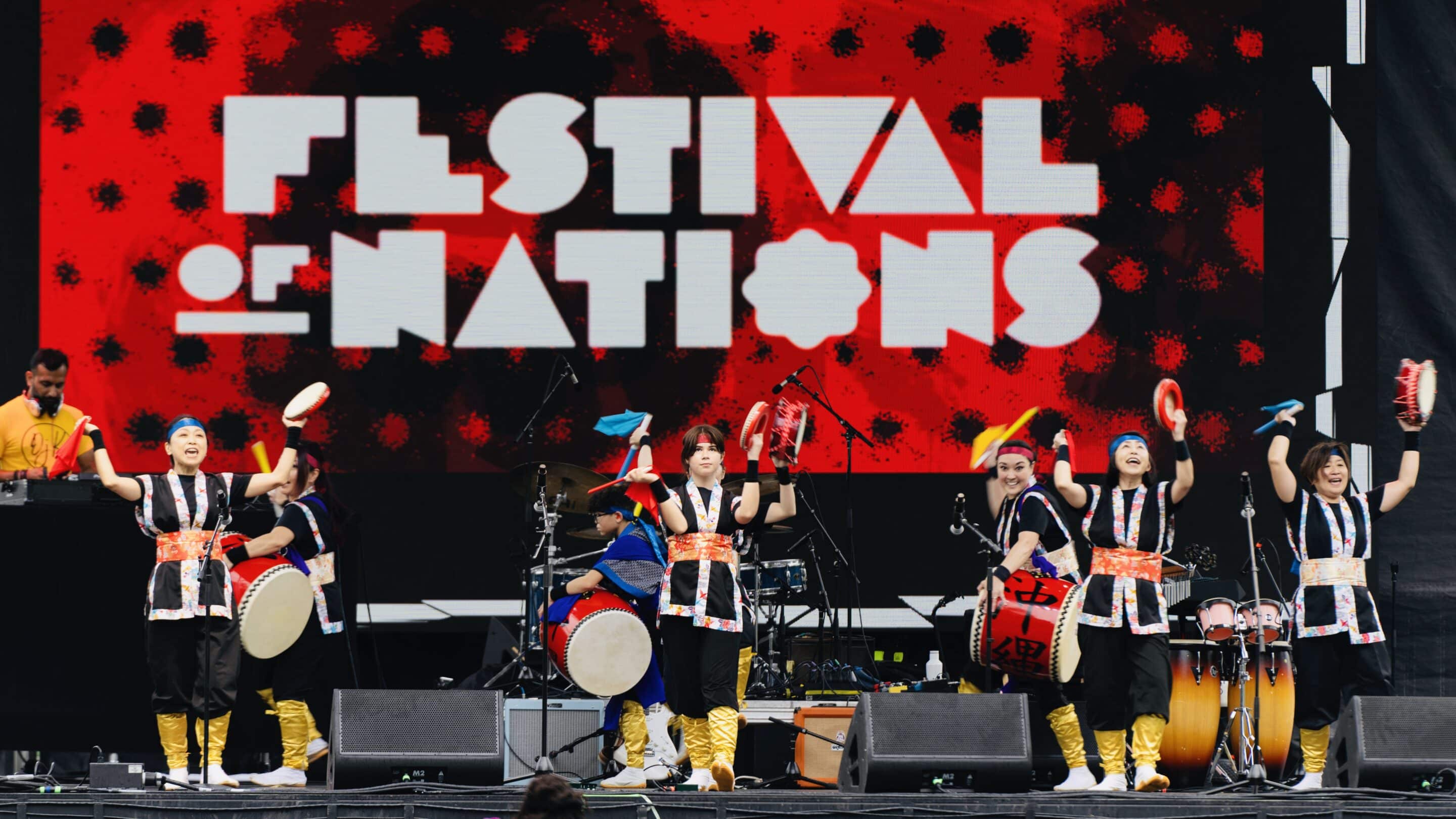 Performers in black costumes with orange sashes and yellow leg wraps playing drums before a red Festival of Nations sign.