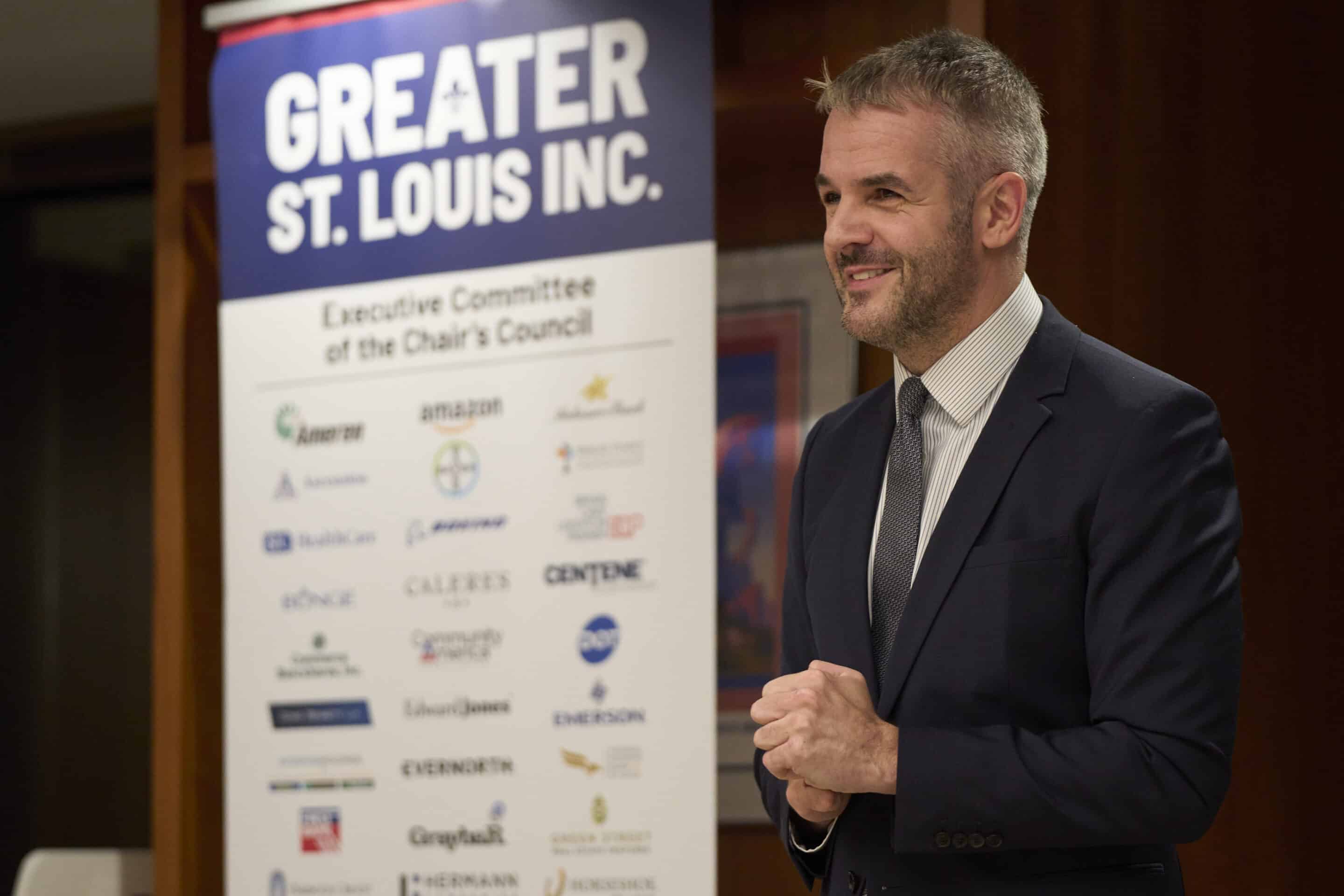 Smiling man in a navy suit with clasped hands speaking beside a "Greater St. Louis Inc." sponsorship banner