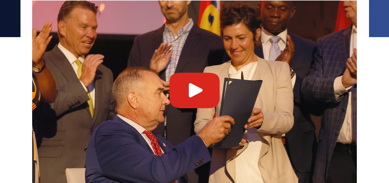 Seated man in blue suit hands a document to a smiling woman on stage as colleagues applaud; red play icon overlay