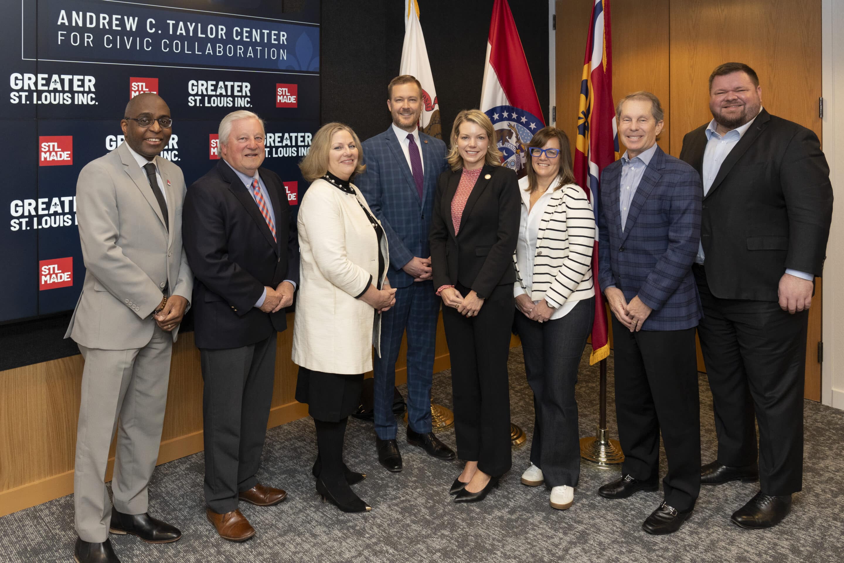 Eight business-dressed people posing in a row before Greater St. Louis signage and flags.