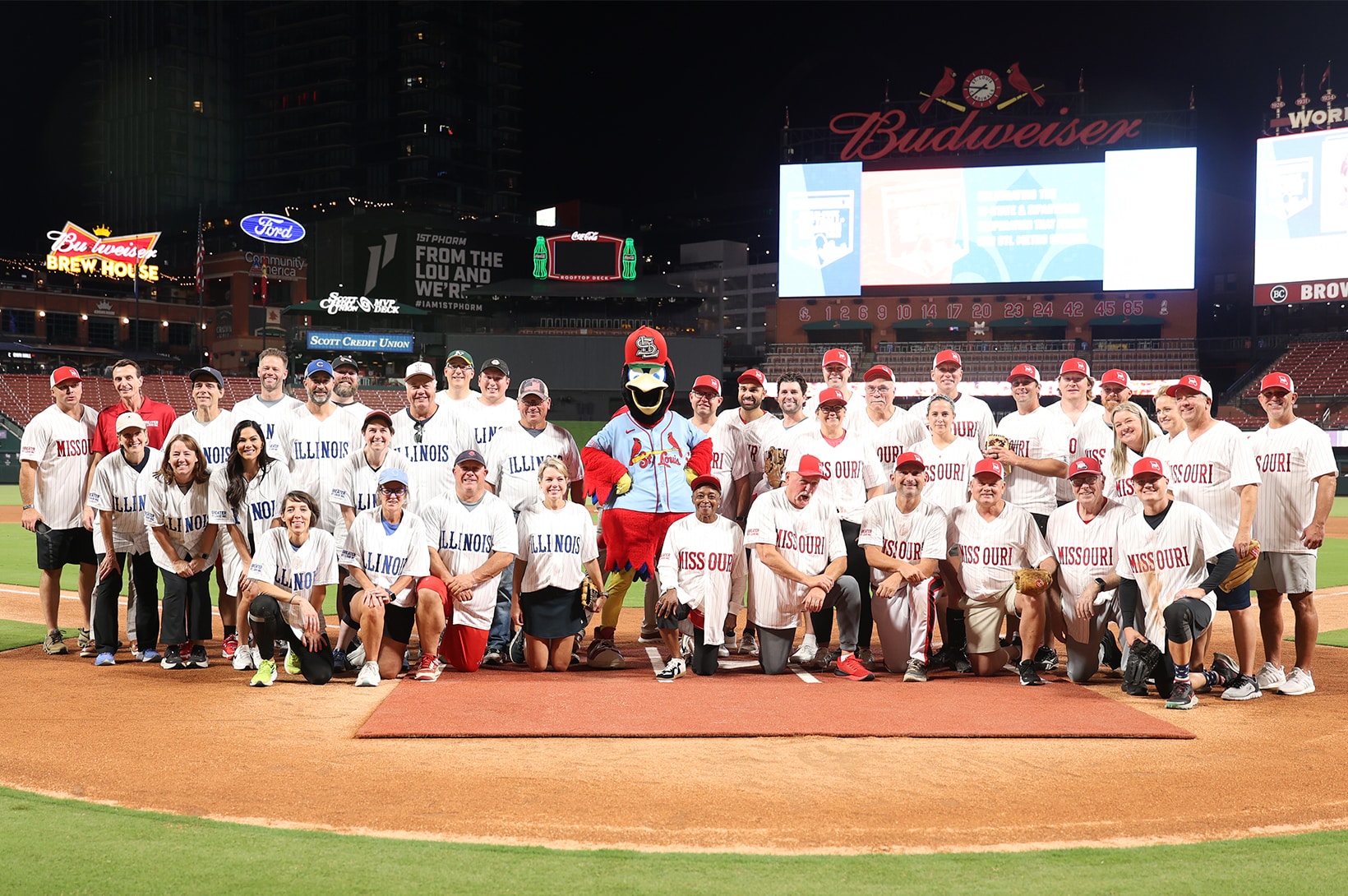 2025 Bi-State Softball Showdown – Group Photo – newsroom Large mixed-age group in Illinois and Missouri baseball jerseys posing with a Cardinals mascot on a stadium field at night.