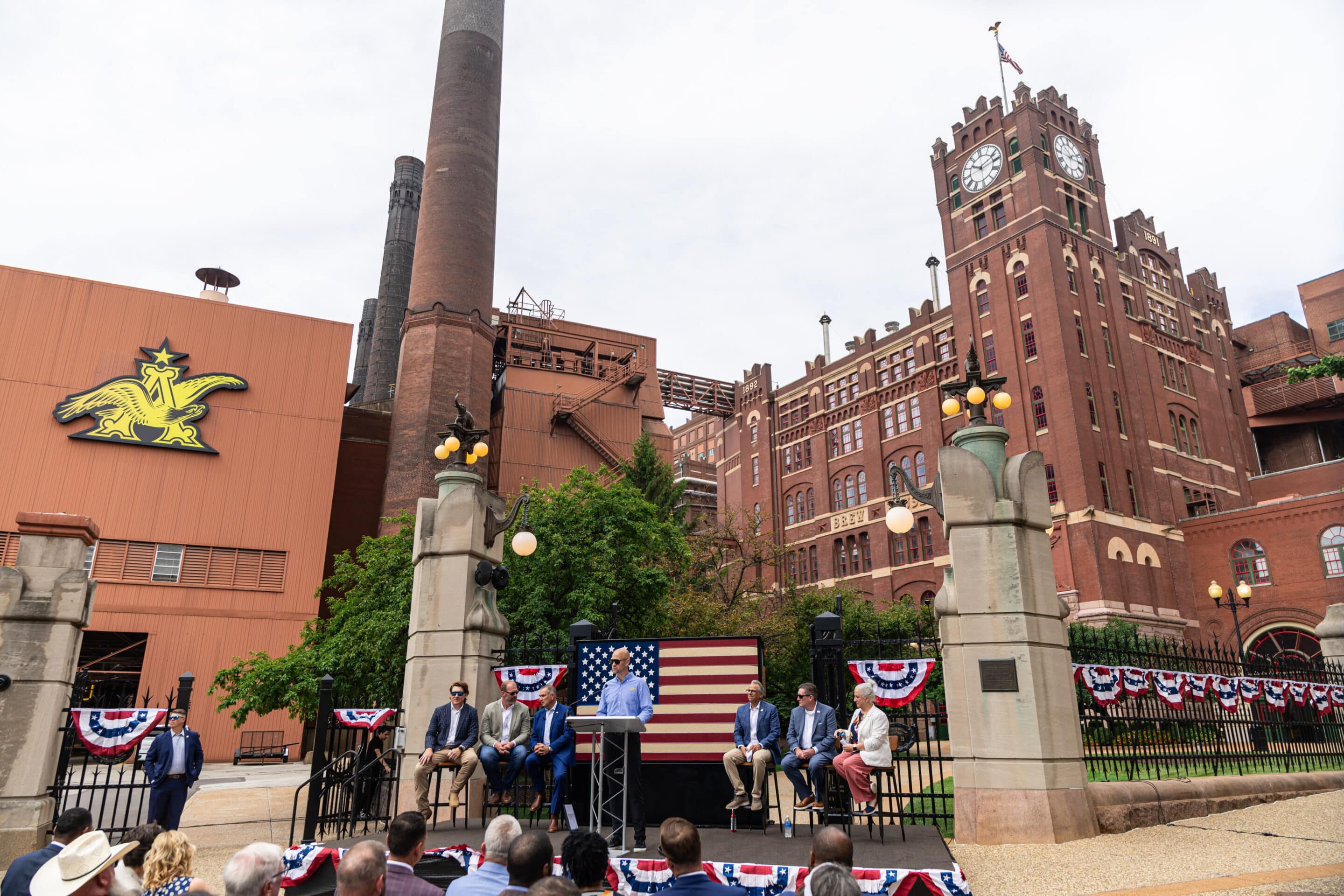 AB-American Beer Act-July 2025 – Newsroom Speaker at a podium with seated panel in front of a brick building with clock tower and American flag backdrop.