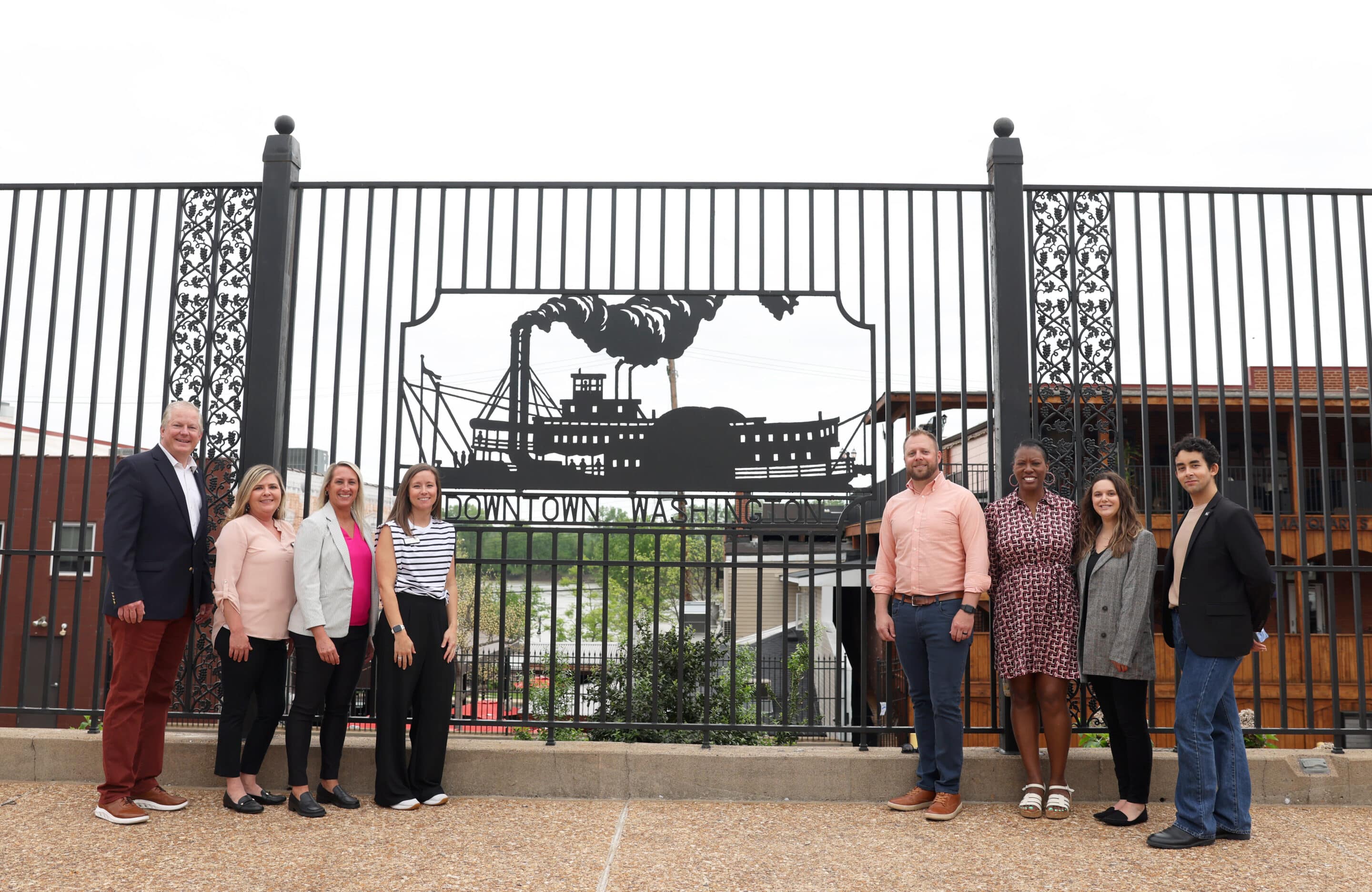Nine people stand in a row in front of an ornamental iron fence featuring a steamboat cutout and a Downtown Washington sign.