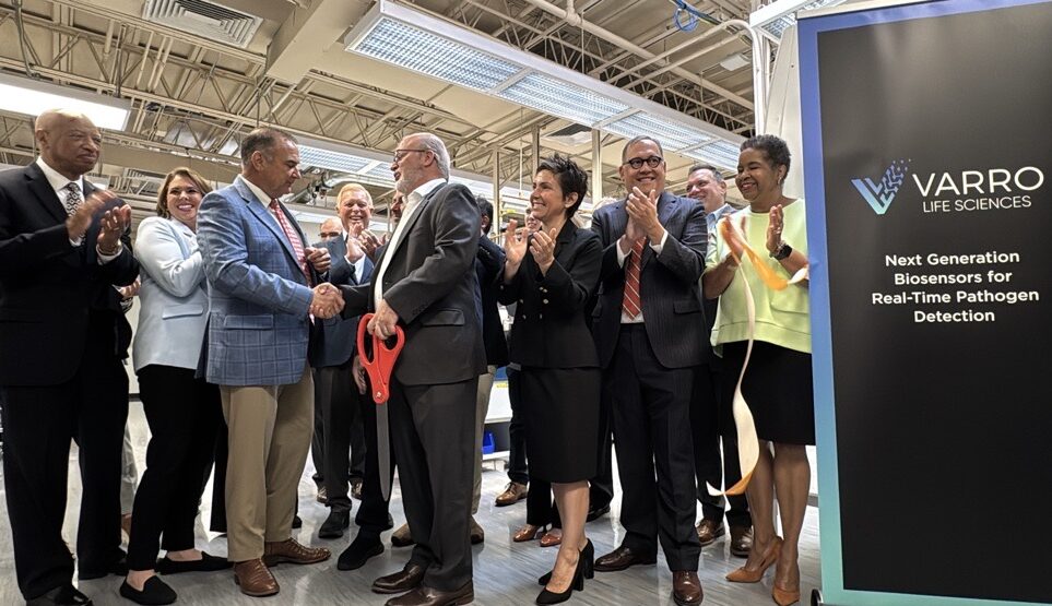 Ribbon-cutting at Varro Life Sciences: man with large red scissors shakes hands as officials applaud by a Varro sign.