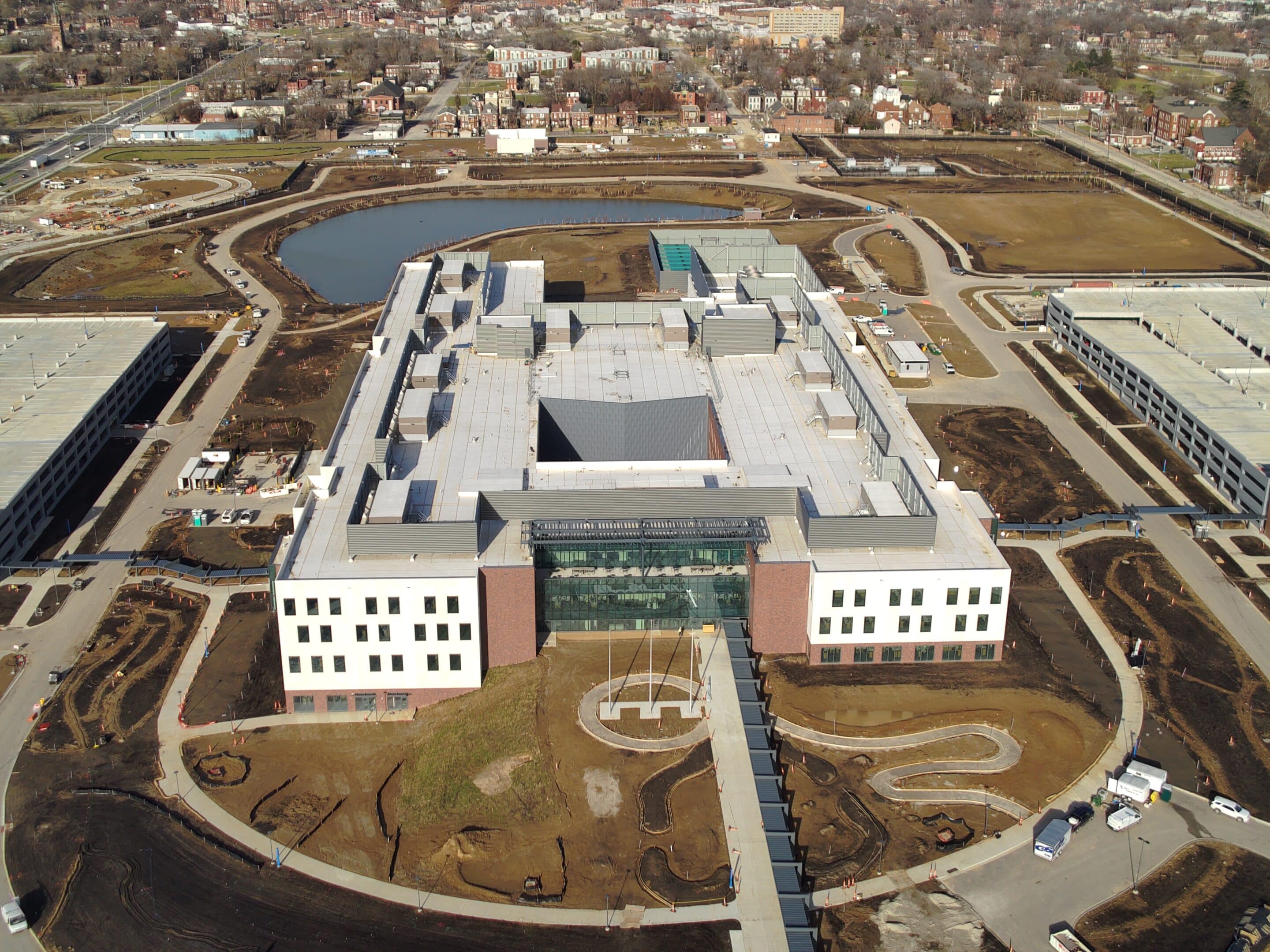 Aerial view of a large rectangular building with a glass central entrance, flanked by parking structures and a pond behind.