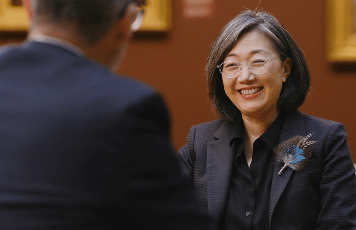 Smiling woman with short hair and round glasses wearing a dark blazer with a blue feather brooch, talking to a blurred man in an art gallery