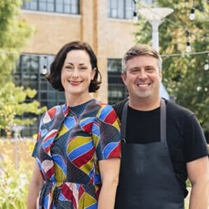 Smiling woman in a colorful patterned dress and man in a black shirt with apron standing outdoors near a brick building.