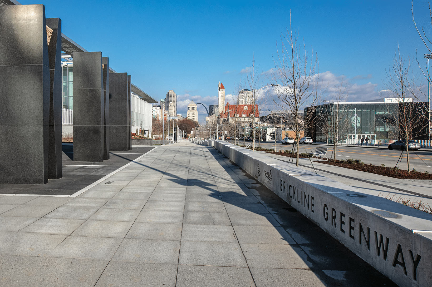 Urban walkway with tall dark stone pillars and concrete bench reading Brickline Greenway, city skyline and arch beyond.