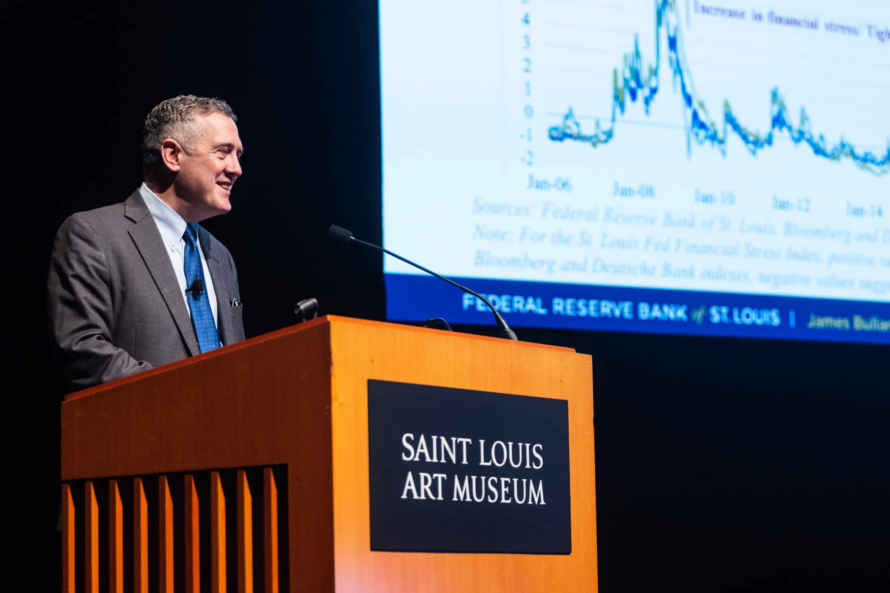 Man in suit speaking at a podium labeled Saint Louis Art Museum with a financial chart projected behind him.