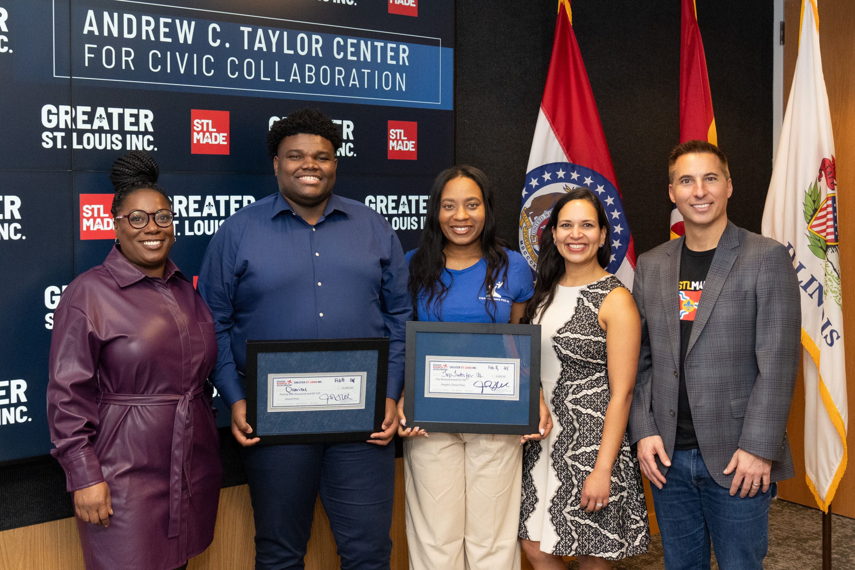 Five people standing side-by-side in front of a civic collaboration backdrop and flags, two holding framed awards and smiling.