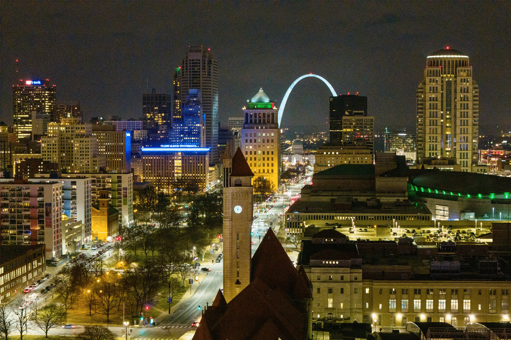 Nighttime downtown skyline with illuminated Gateway Arch, a prominent clock tower with red roof, and lit office buildings.