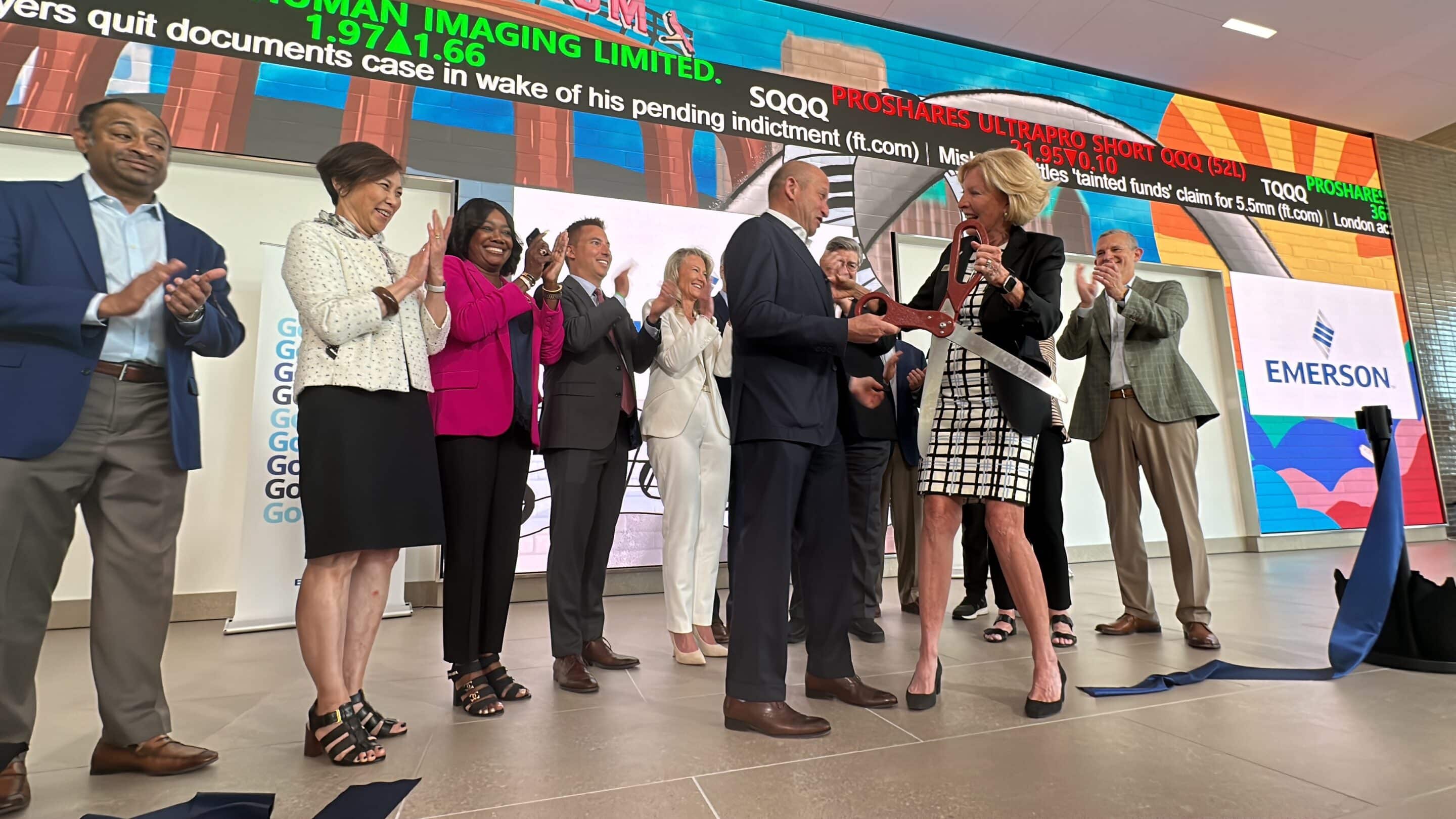 Woman uses oversized scissors to cut a blue ribbon while a group of colleagues applauds in front of a digital Emerson sign.