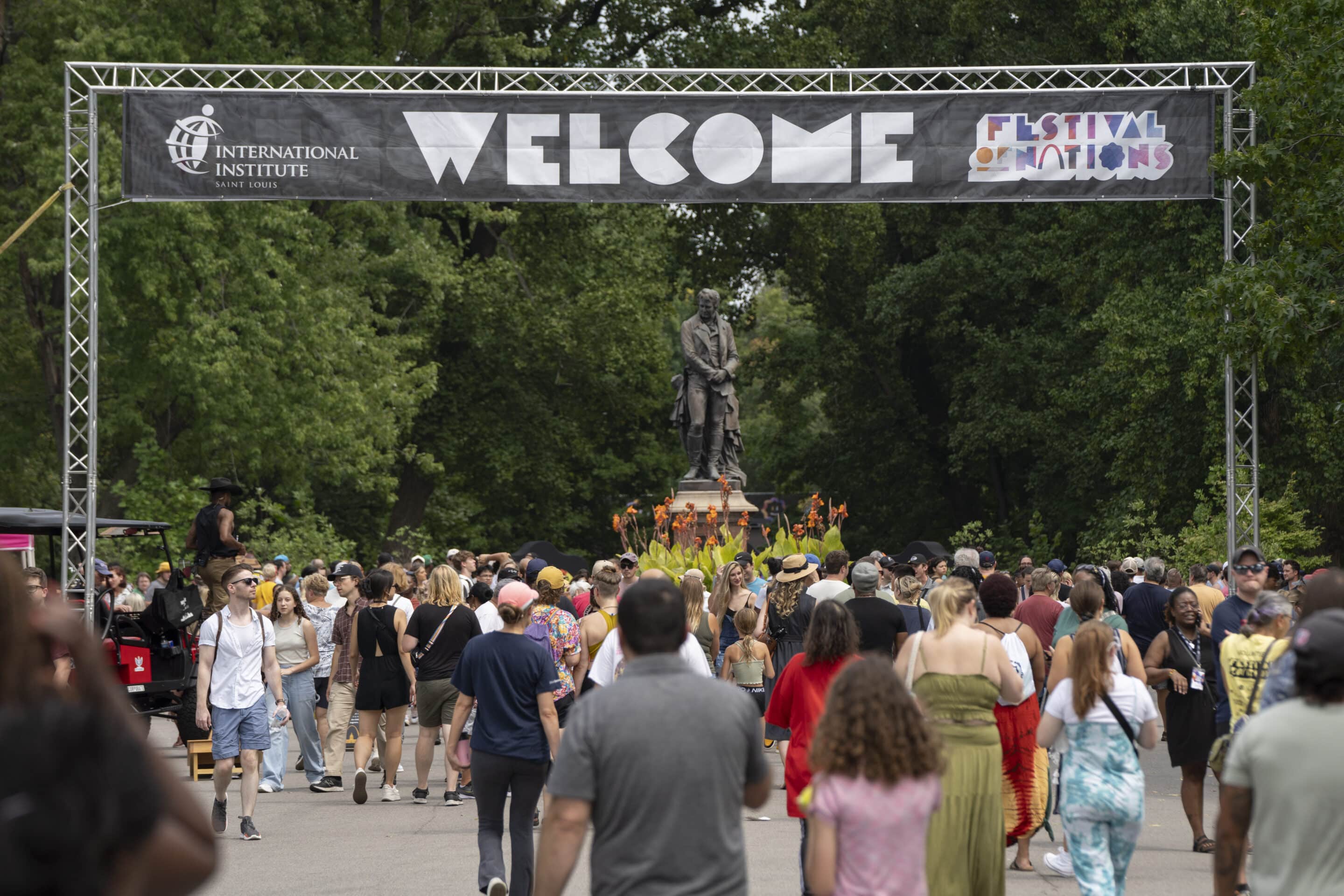 Crowd walking under a large WELCOME banner at an outdoor festival, a statue and trees visible ahead.