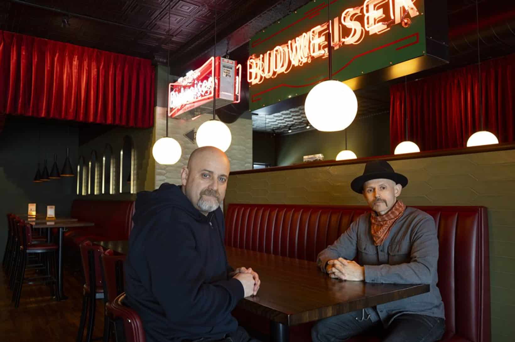 Two men sitting at a red leather booth in a dim bar with a neon Budweiser sign and round globe pendant lights.