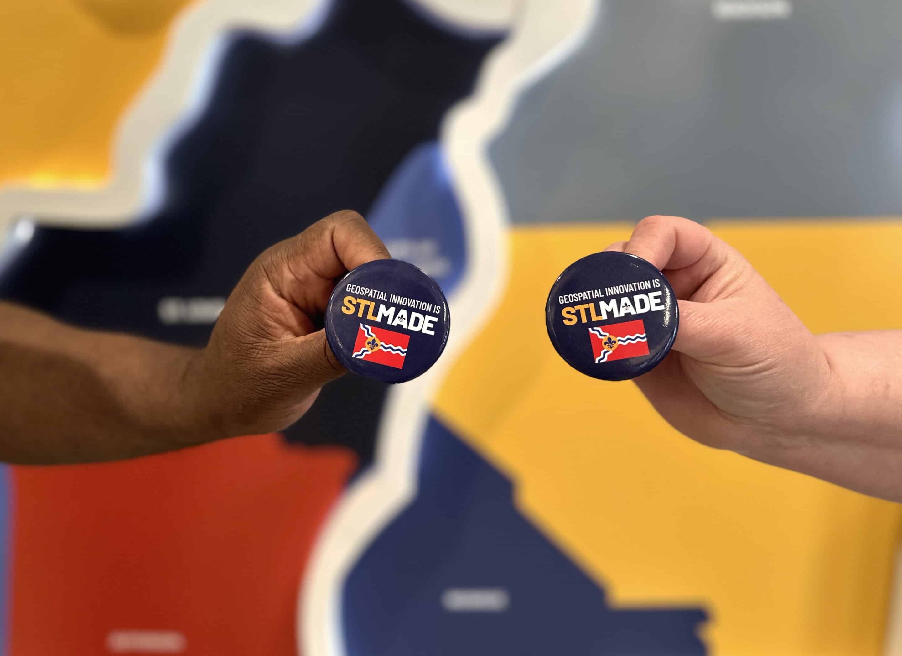 Two hands holding navy buttons reading STL MADE with a small red flag emblem against a colorful map background.