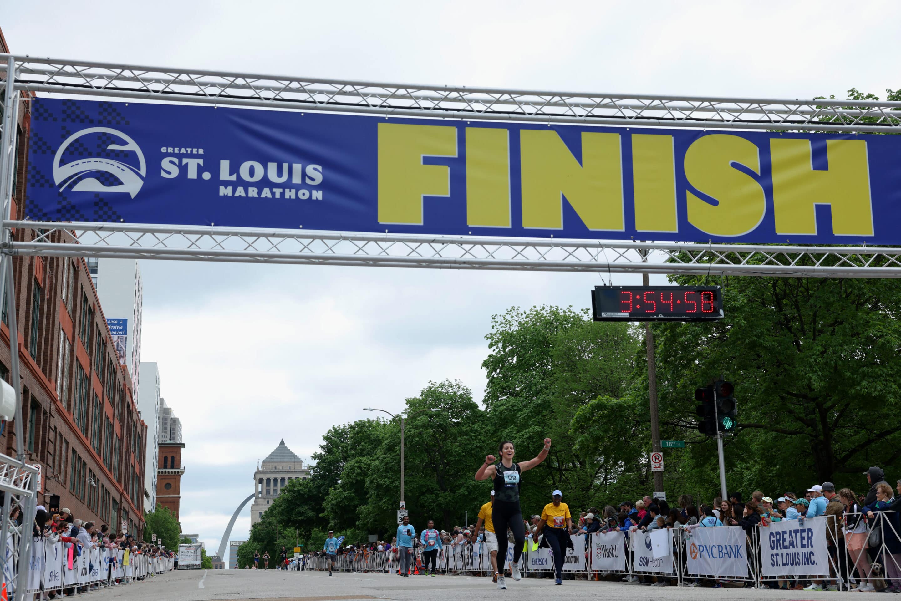 Runner raising arms crossing finish line under large FINISH banner at St. Louis Marathon with Arch in background.