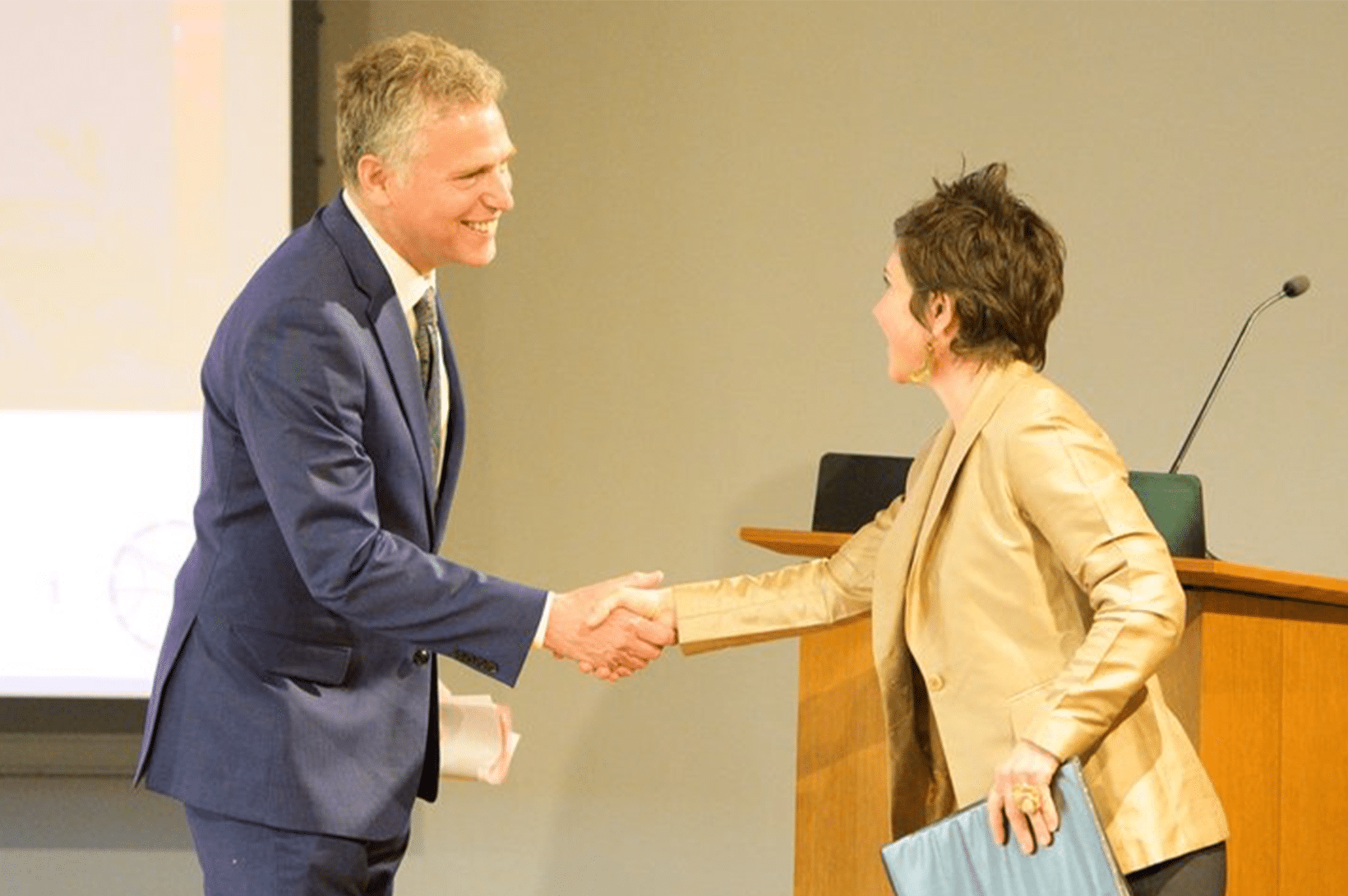 gsl_news_brain_power.png Man in a blue suit and woman in a tan blazer smile and shake hands across a wooden podium while the woman holds a blue folder