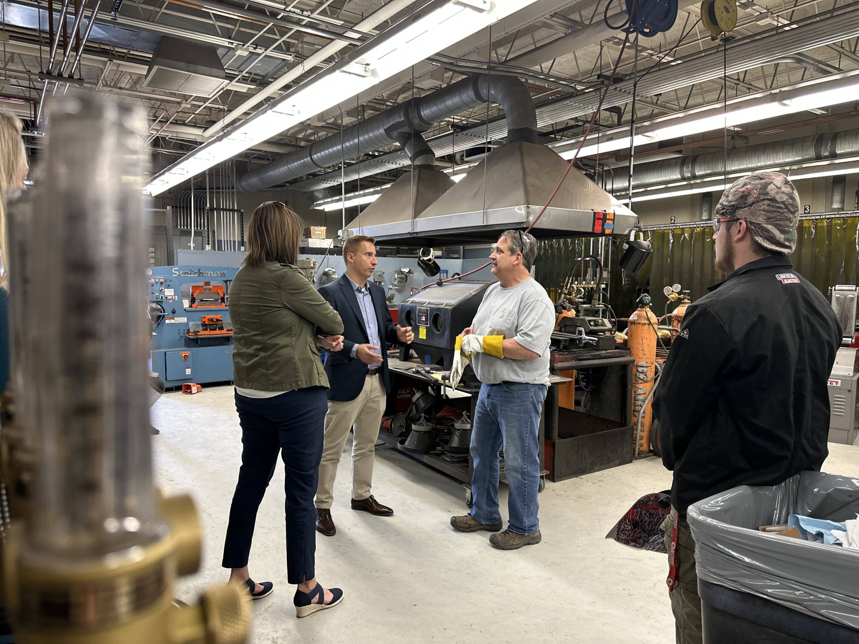 img_2180.jpg Four people standing in a metalworking shop conversing near a welding hood and gas tanks, one man wearing yellow gloves.