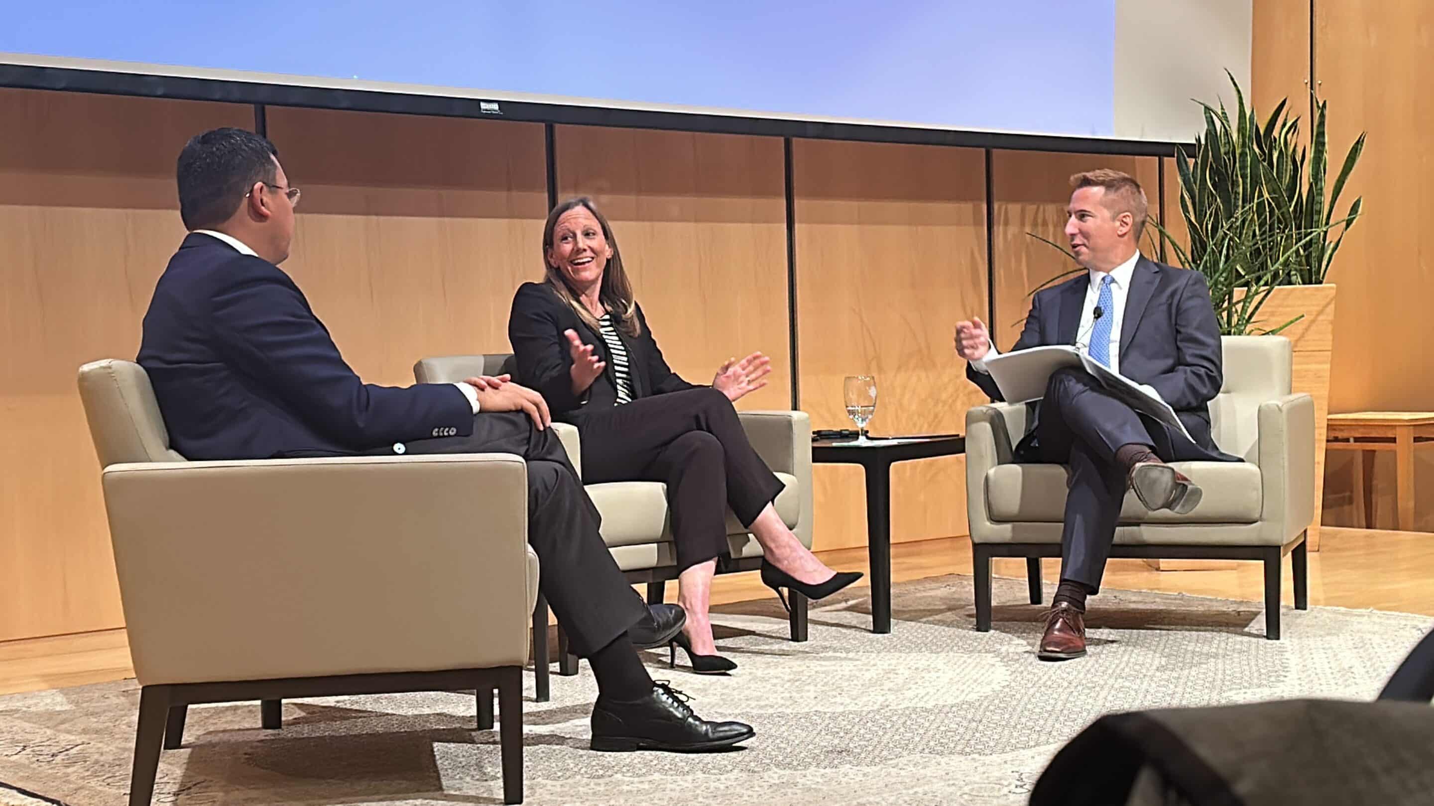 Three panelists seated in armchairs on stage; woman in center gesturing while two men listen, small table with glass.