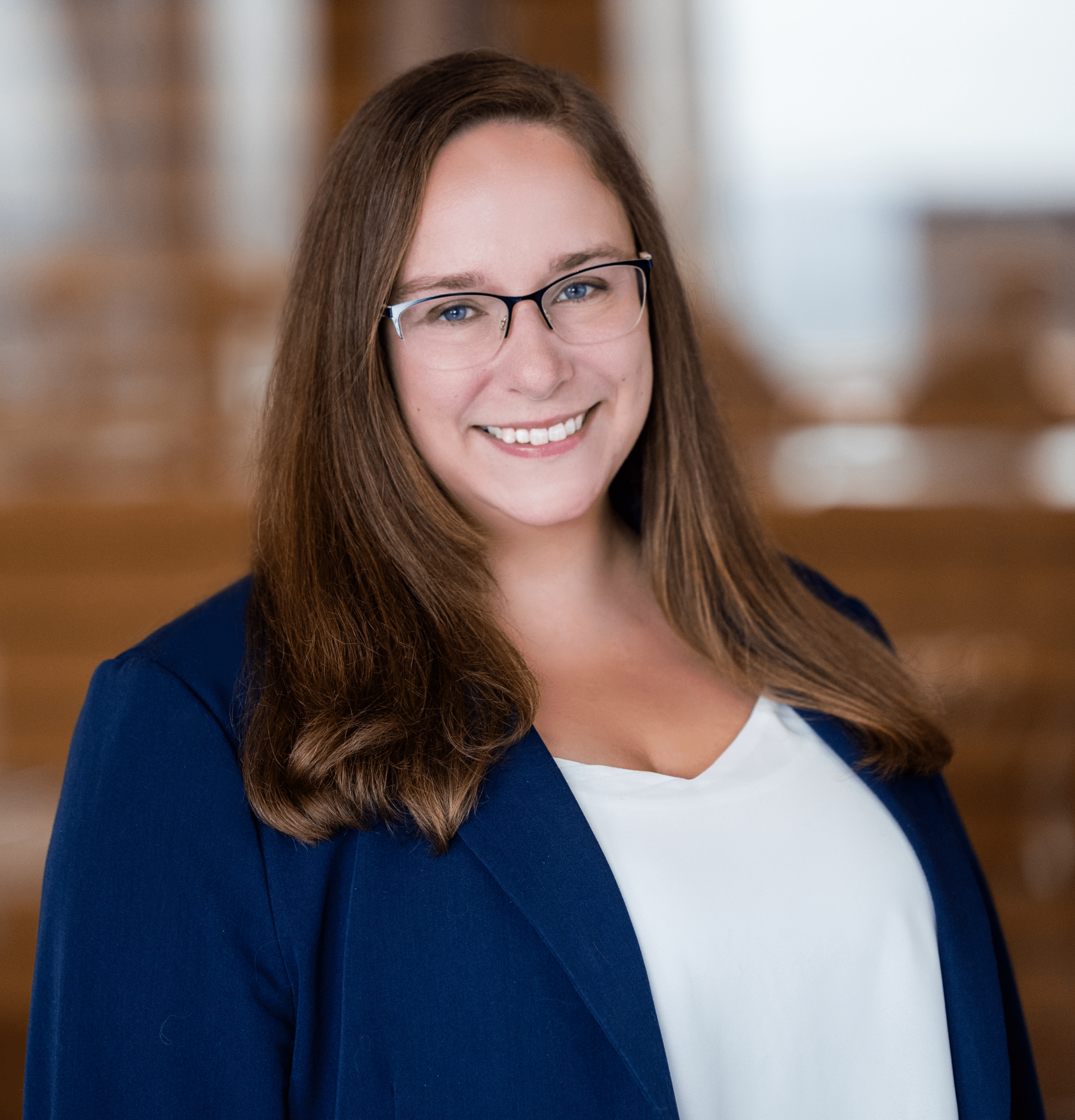 Smiling woman with long brown hair and glasses wearing a navy blazer and white blouse against a blurred indoor background.