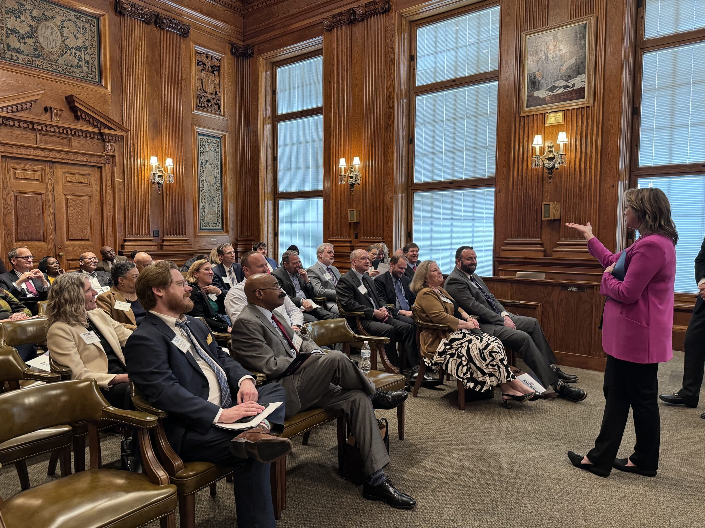 Woman in pink blazer speaking to a seated audience in a wood-paneled meeting room with tall windows.
