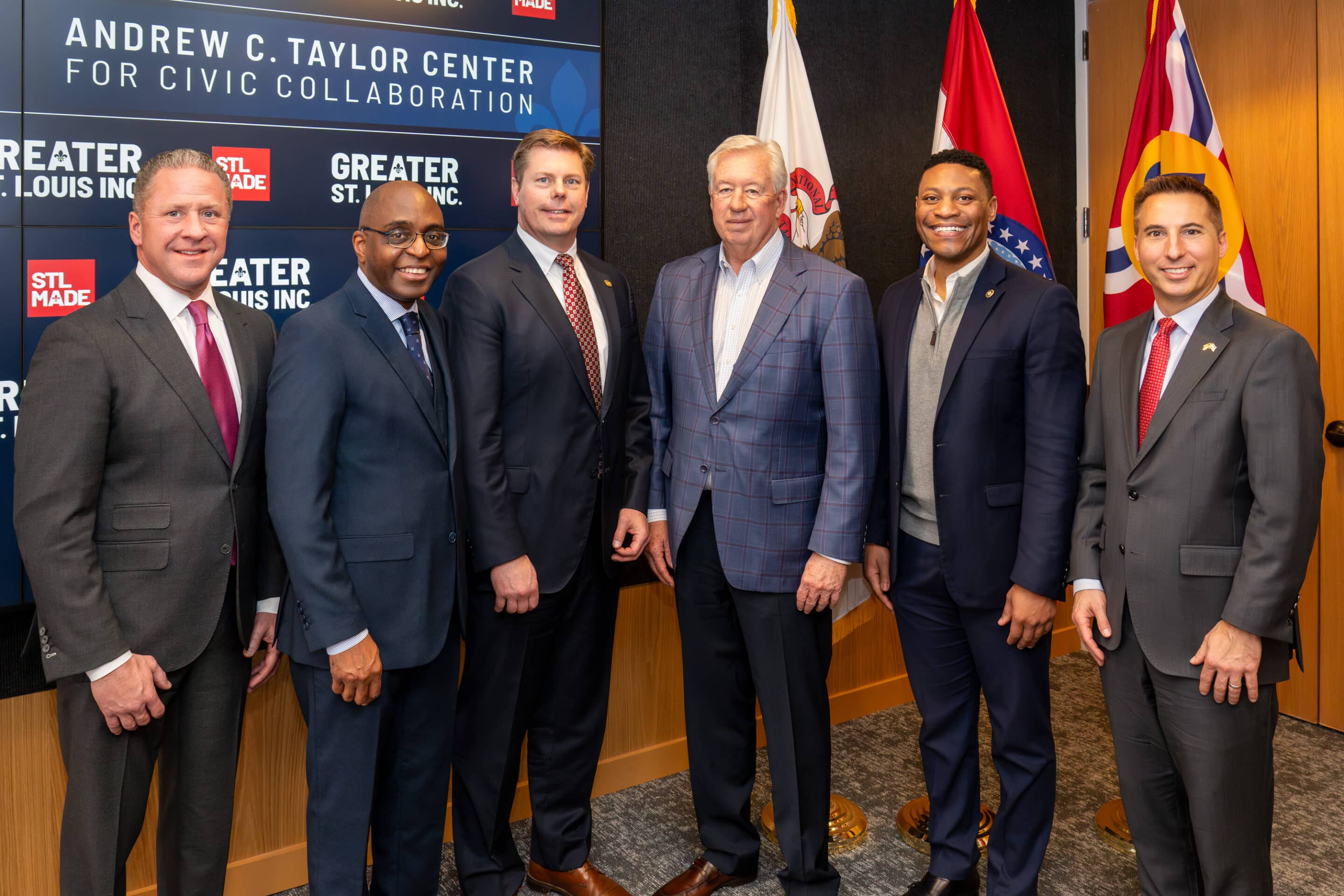 Six men in suits standing side-by-side, smiling in front of a sign reading Andrew C. Taylor Center and flags.