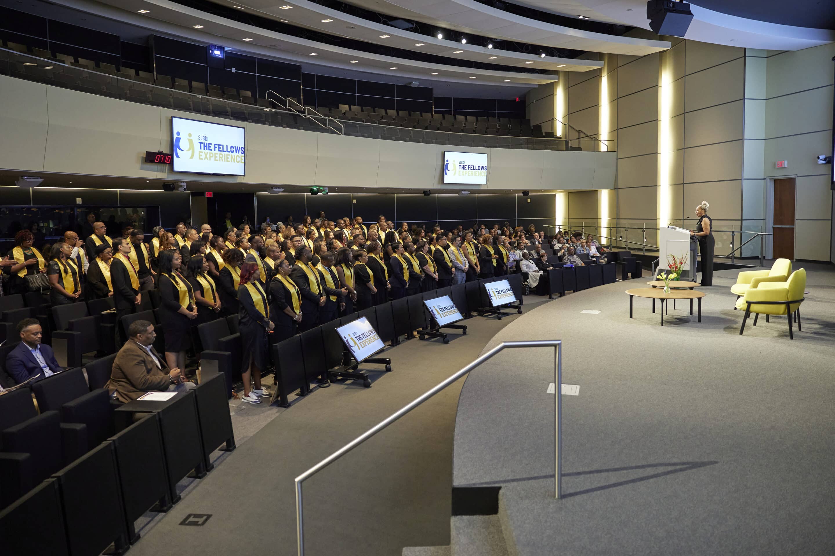 Large group of people wearing yellow stoles stand in an auditorium facing a woman speaking at a podium on stage.
