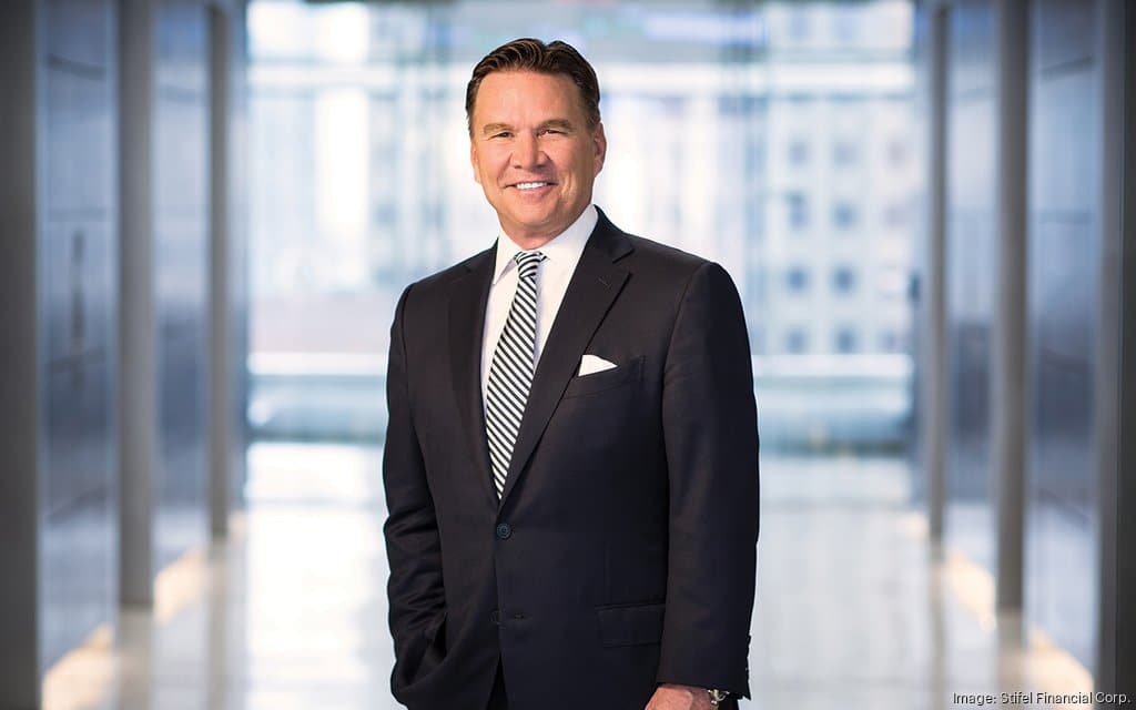 Smiling middle-aged man in a dark suit and striped tie standing in a bright modern office corridor.