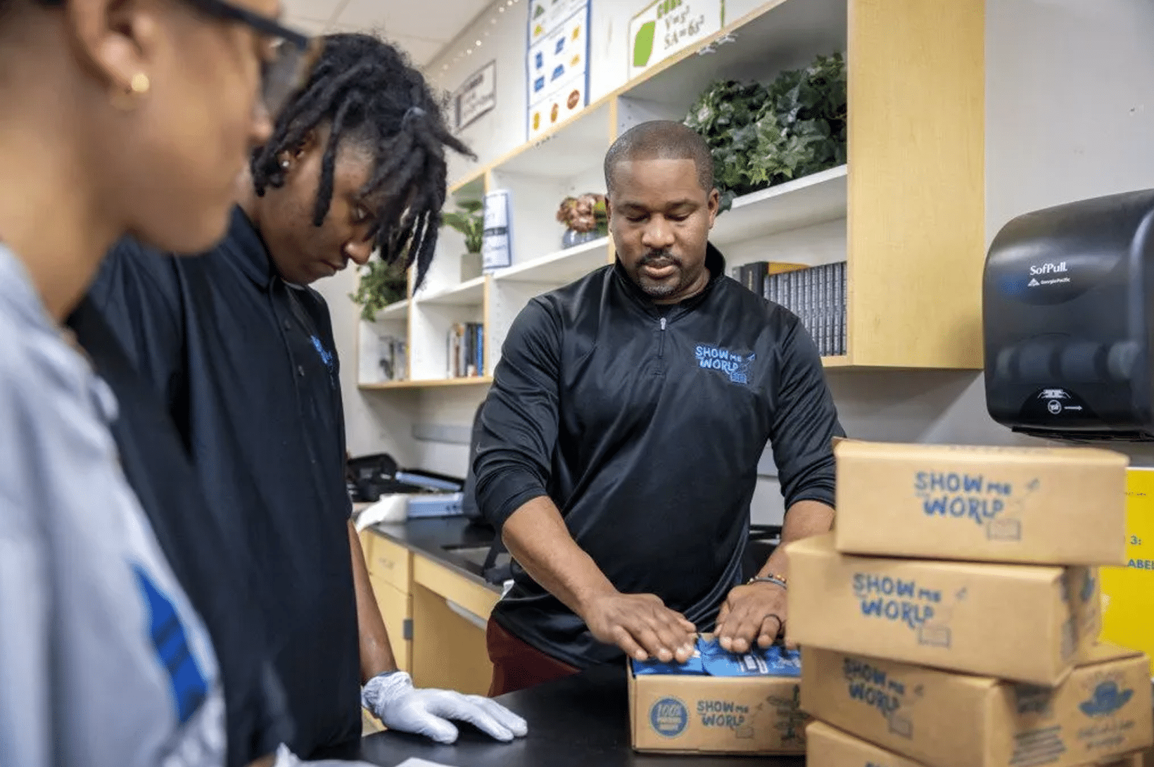 Man in black Show Me the World shirt pressing down on a packaged box as two students watch beside a stack of labeled boxes.