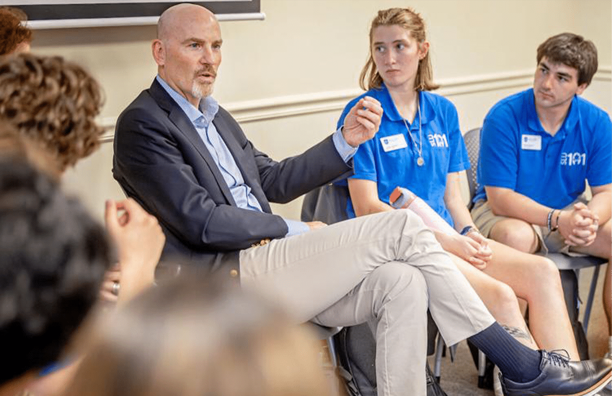 slu_leader_takes_helm.png Bald man in a blazer and khaki pants speaking to a circle of students, with two teens in blue polo shirts listening