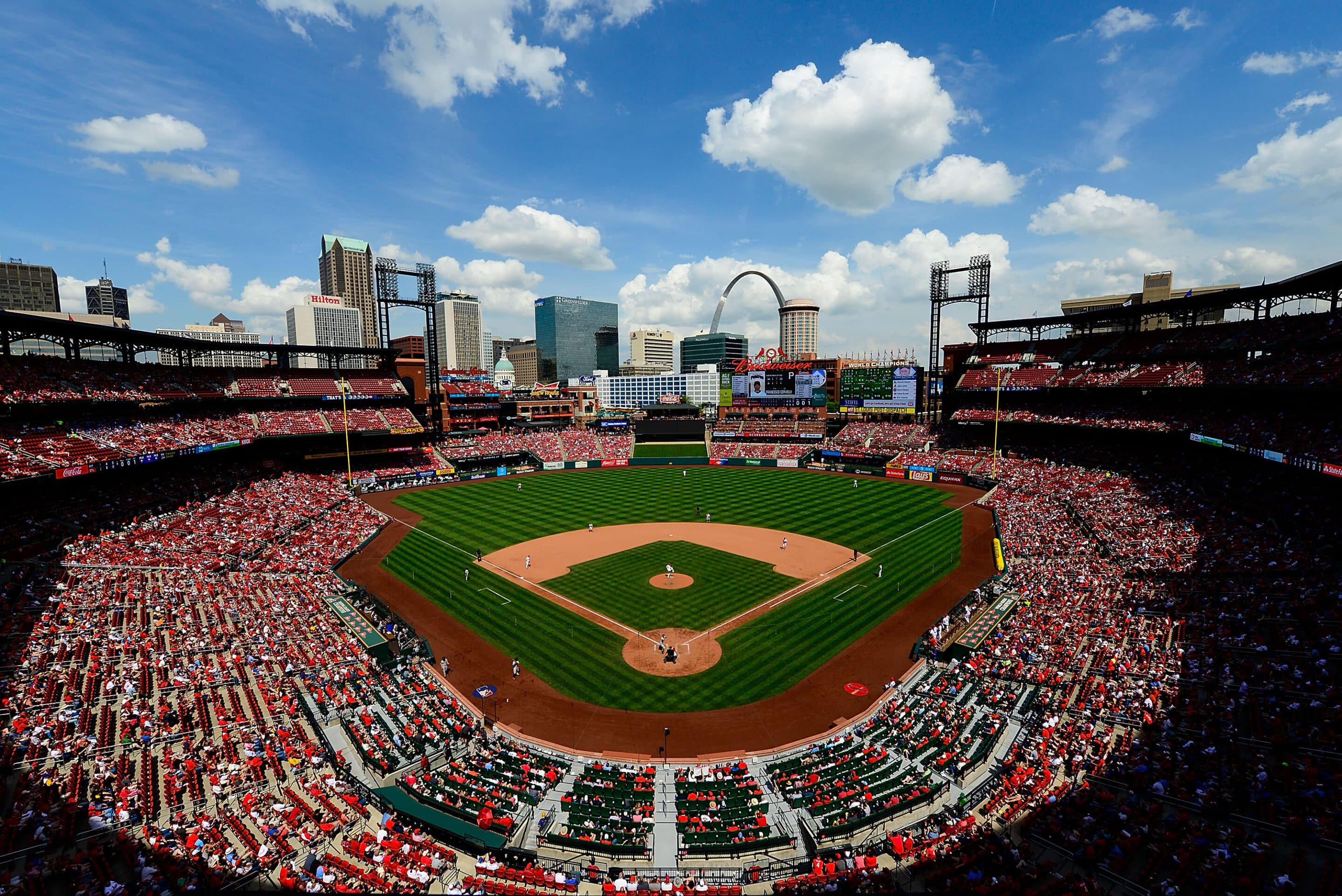 Panoramic view of a packed baseball stadium with green infield and the Gateway Arch in the background on a sunny day.