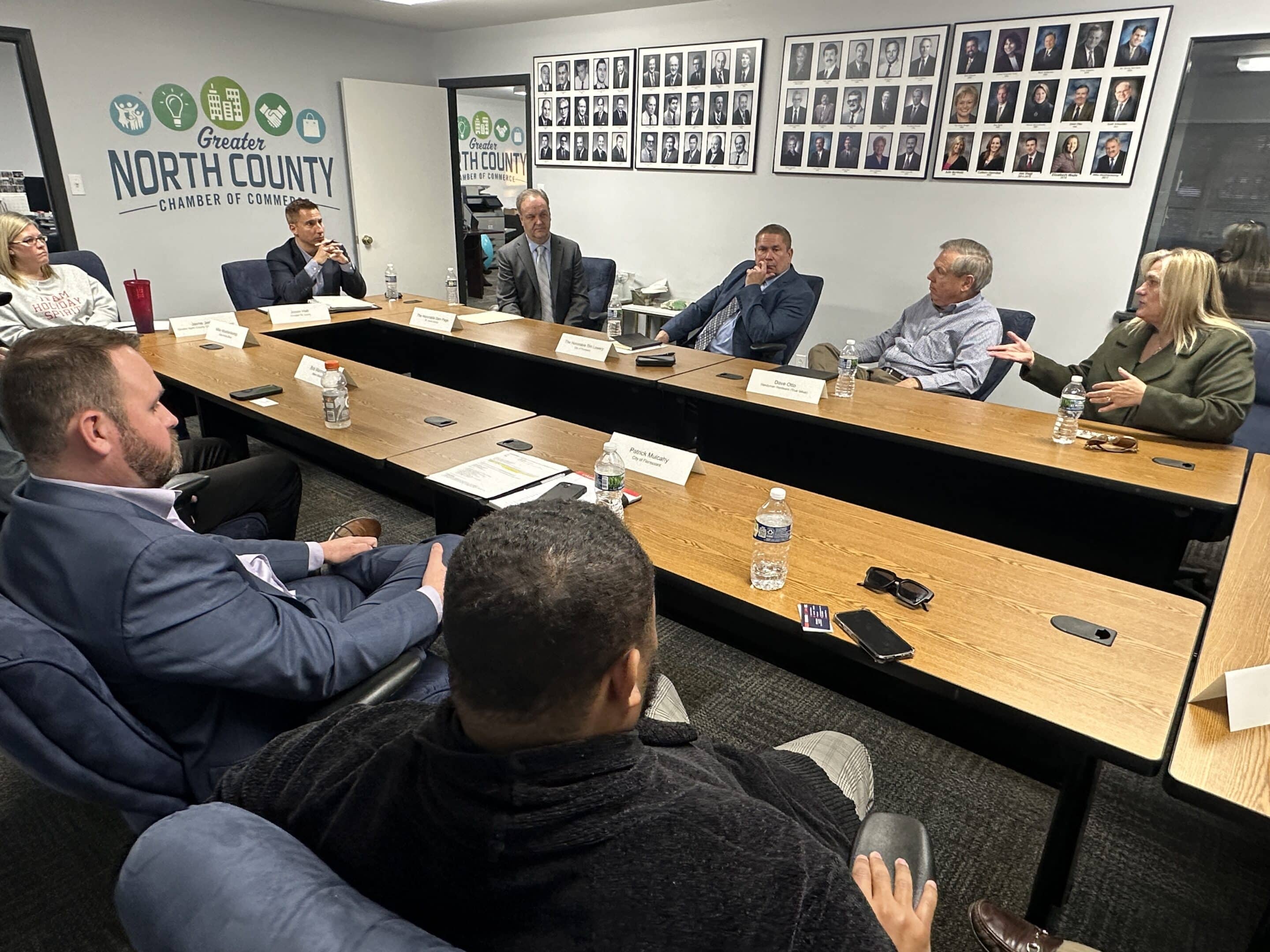 Business meeting around a conference table with several people discussing under Greater North County Chamber of Commerce logo.