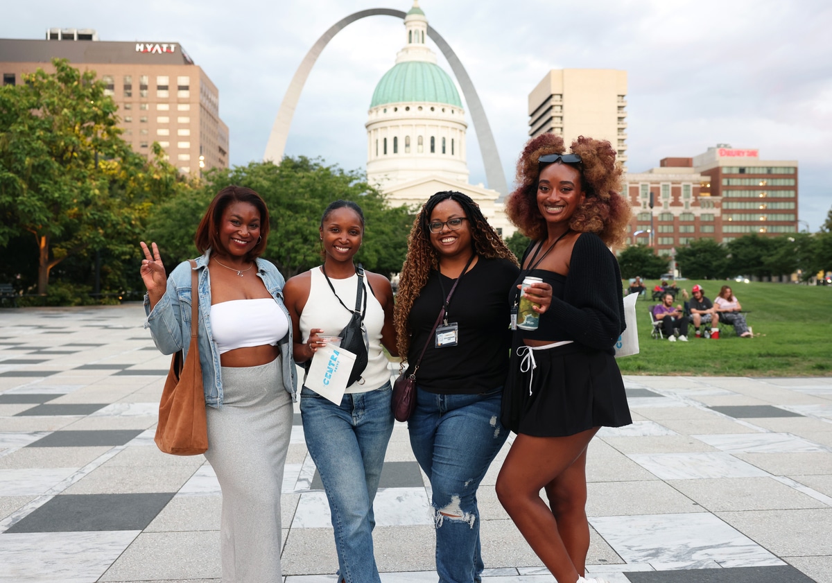 Long row of varied beer tap handles mounted above a stainless-steel drip tray, with wooden shelves of bottles and glassware on a brick back wall.