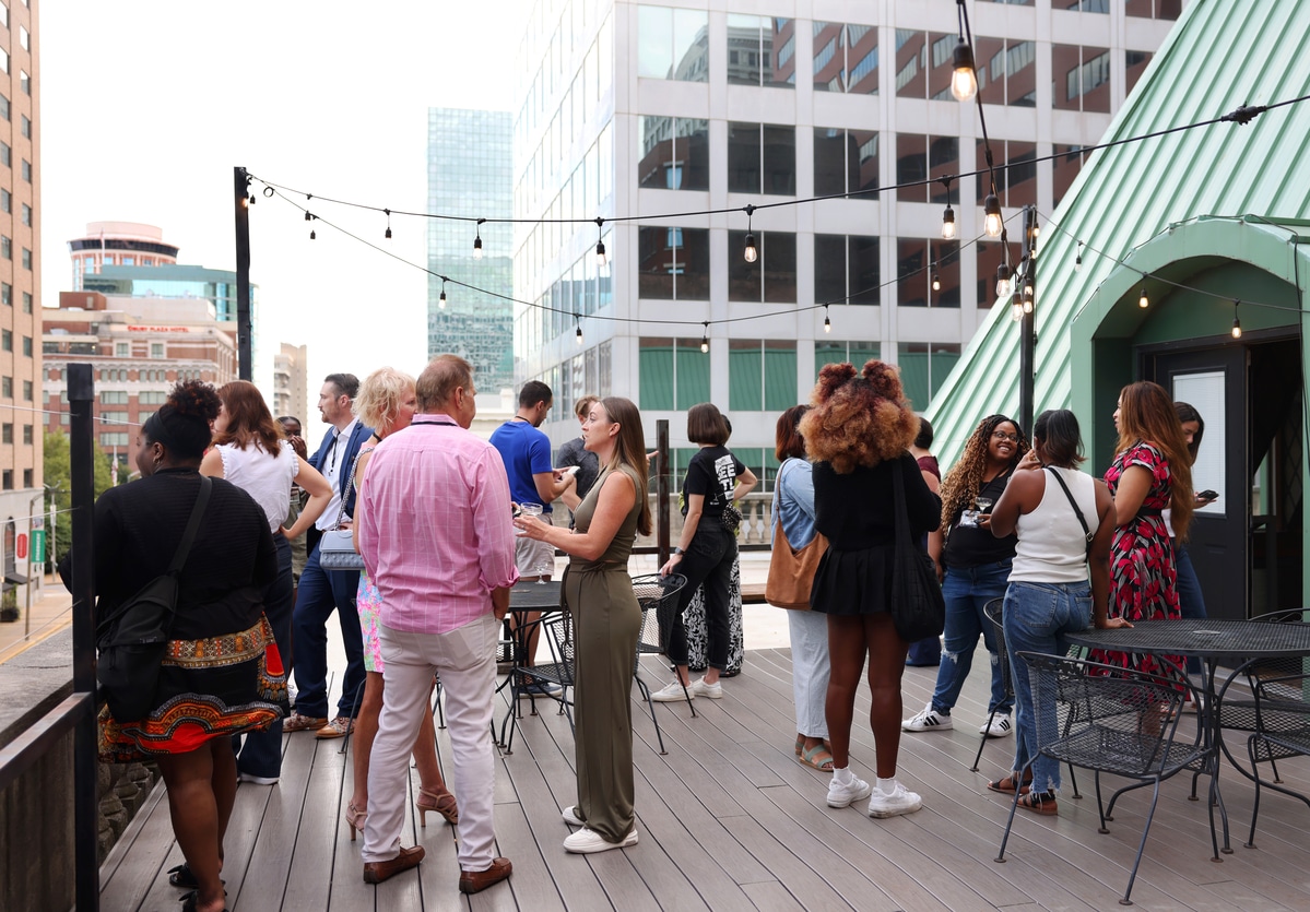 Rooftop social gathering of a diverse group chatting under string lights with city buildings in the background.