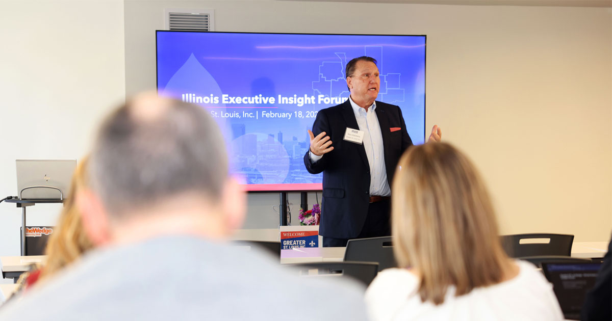 Man in suit speaking in front of a presentation screen that reads "Illinois Executive Insight Forum," with audience heads in foreground