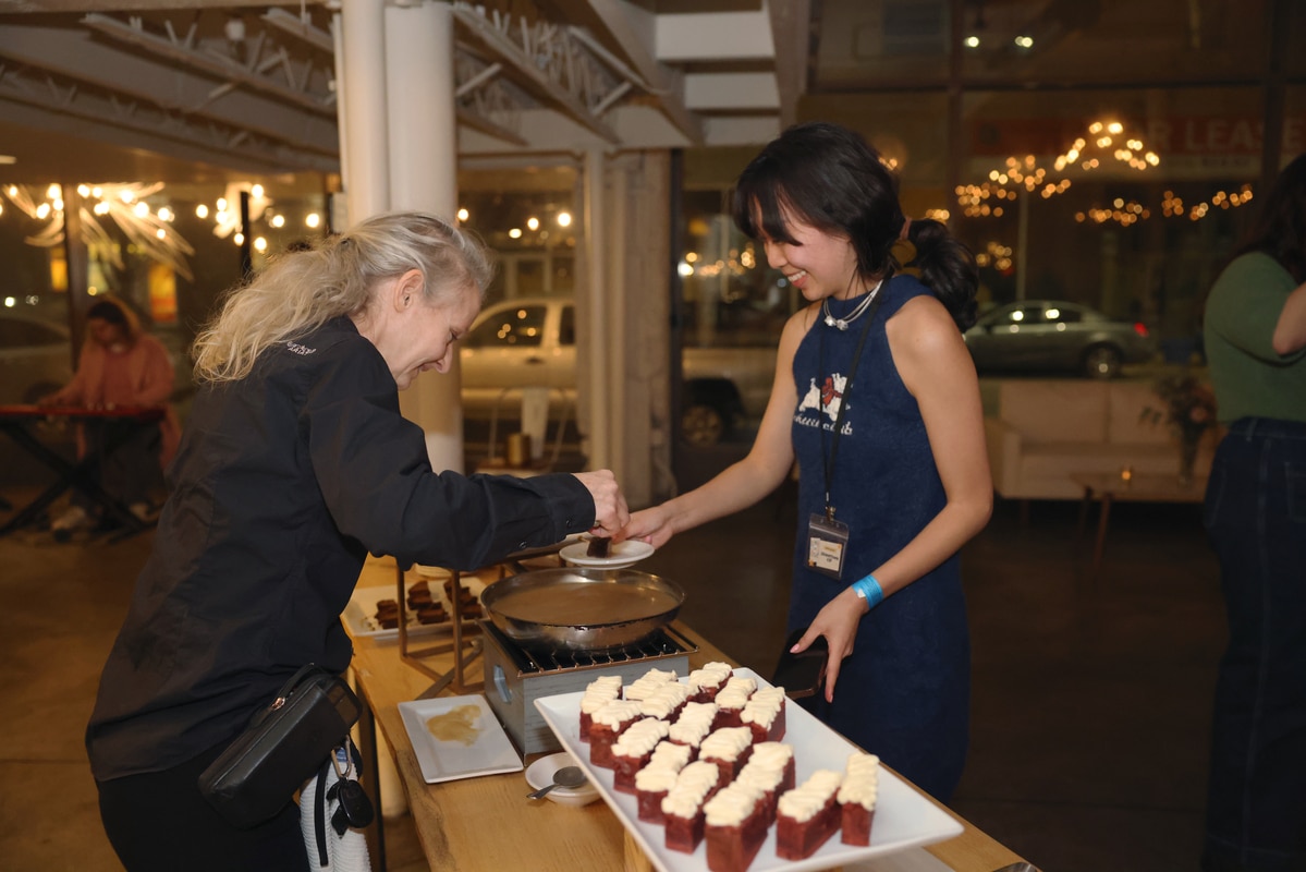 Woman in black serving a frosted cake slice to a smiling woman in a blue dress beside a tray of small cake pieces.