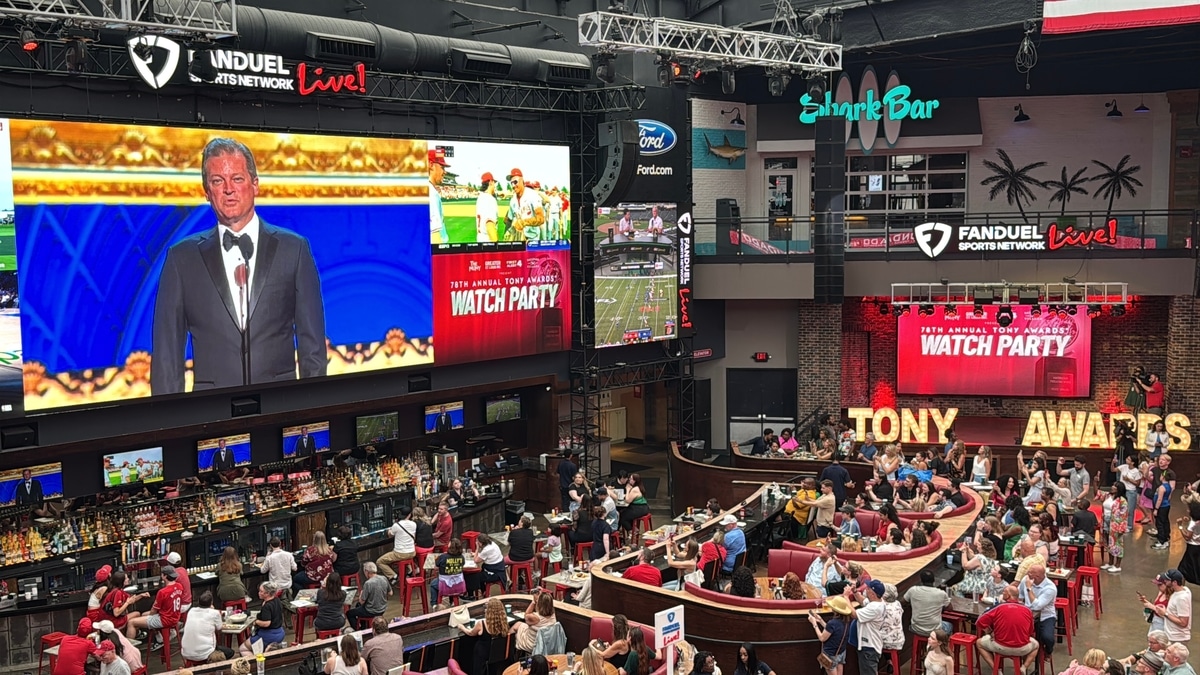 The Muny Tony Awards Watch Party Ballpark Village 2025 9-1200×675-128554e Crowded indoor watch-party bar with a huge screen showing a tuxedoed presenter and lit TONY AWARDS letters.
