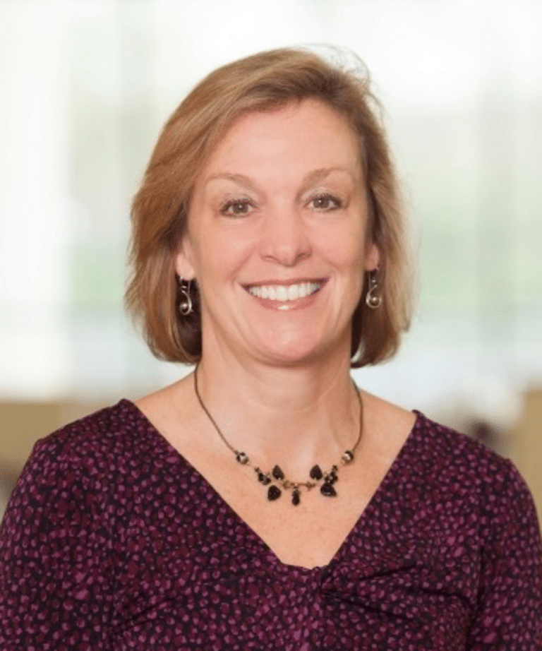 Smiling middle-aged woman with short light brown hair wearing a purple patterned blouse and a black beaded pendant necklace.