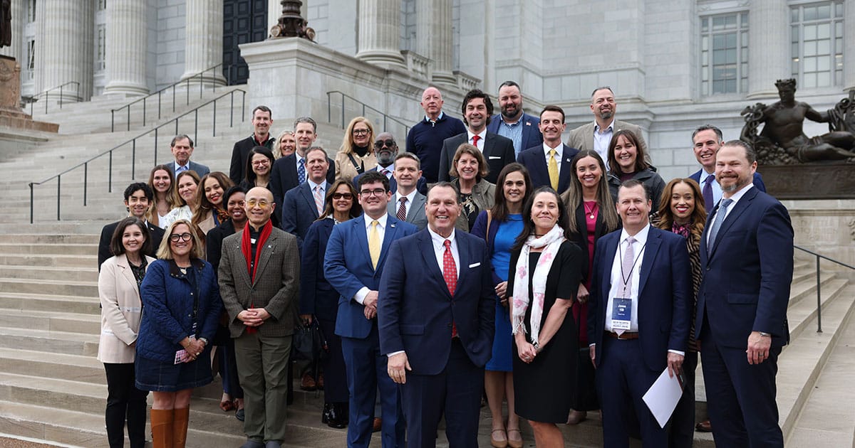 Group of professionally dressed men and women posing on stone steps of a large neoclassical building, smiling toward the camera.