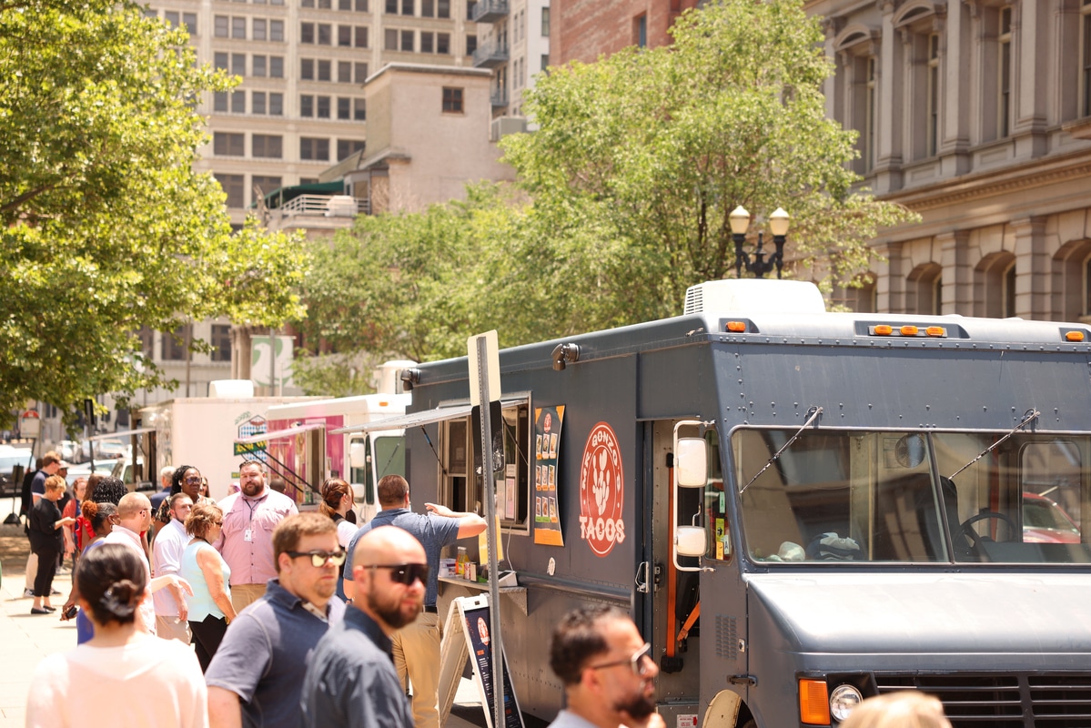 Crowd of people lined up at a dark blue taco food truck parked on a sunny downtown street with trees and tall buildings.