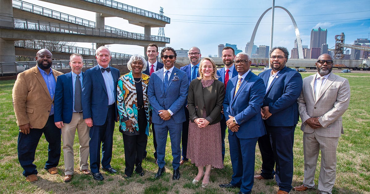 Twelve diverse professionals in business and business-casual attire standing on grass with a city skyline and a large arch monument in the background.