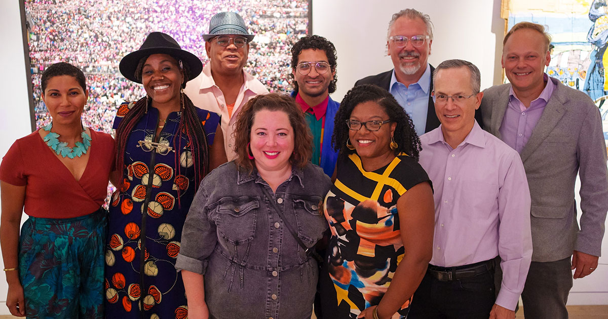 Group of ten smiling adults standing in two rows inside a gallery, wearing colorful clothing and posing in front of artwork.