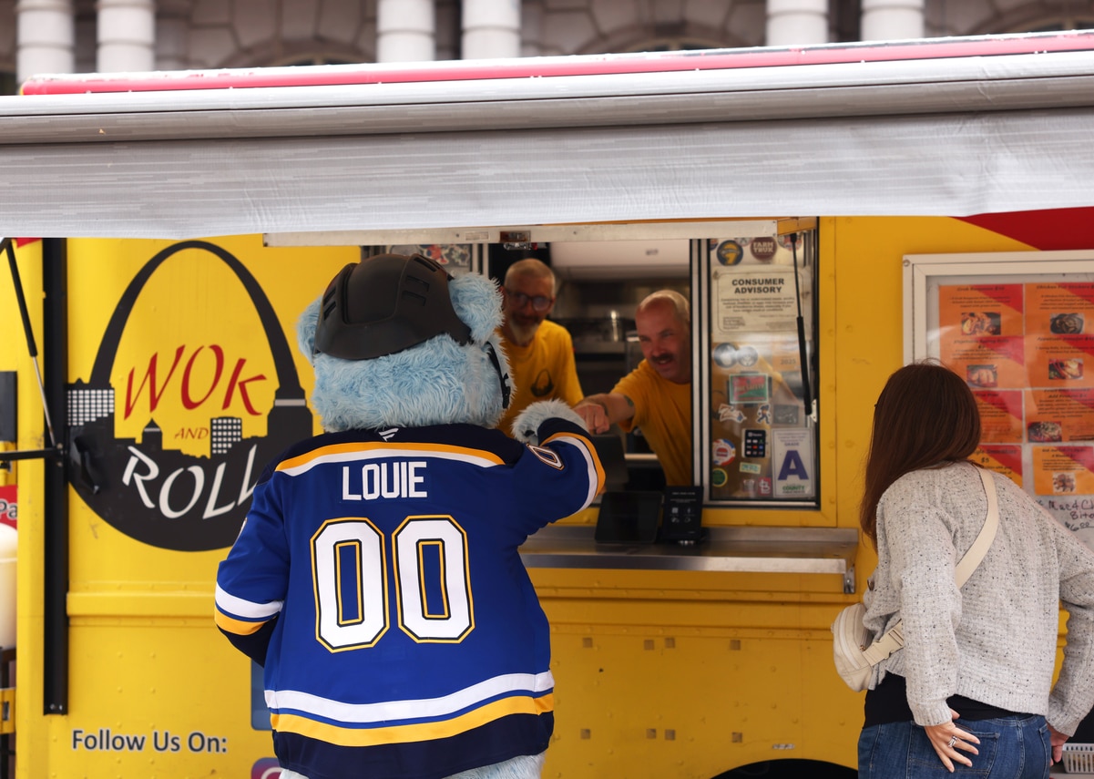 Blue furry mascot in LOUIE 00 hockey jersey and helmet reaches into yellow Wok and Roll food truck as vendors hand an order