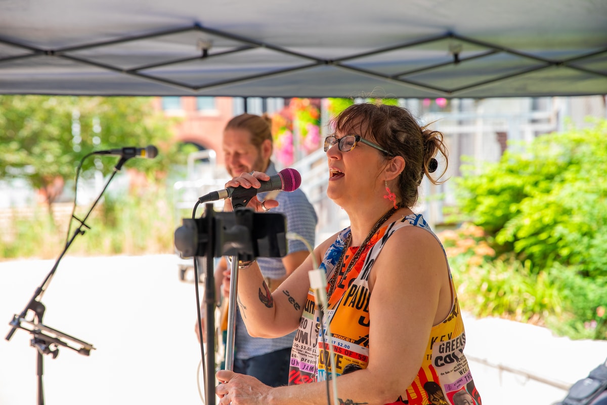 Woman with glasses and flamingo earrings singing into a pink-covered microphone under a canopy, guitarist blurred behind her.