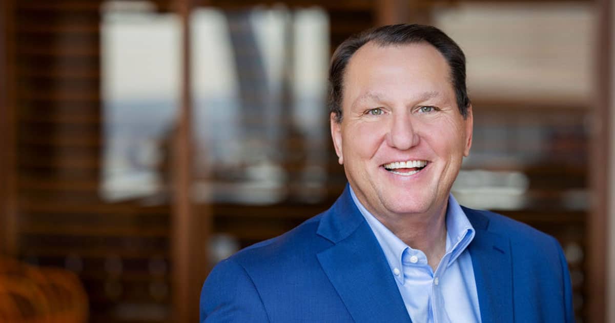 Ron Kitchens in a blue suit and light blue shirt, shown from the chest up against a blurred indoor background.