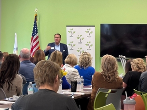 Man in suit speaking to seated audience in a meeting room with US flag, plant-themed banner, and large TV on green wall.