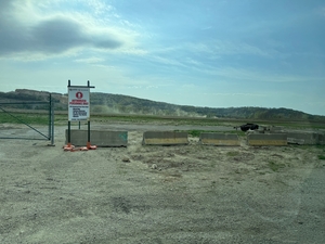 Empty gravel lot with concrete barriers and a warning sign in front of a wide open landscape and low hills under a partly cloudy sky